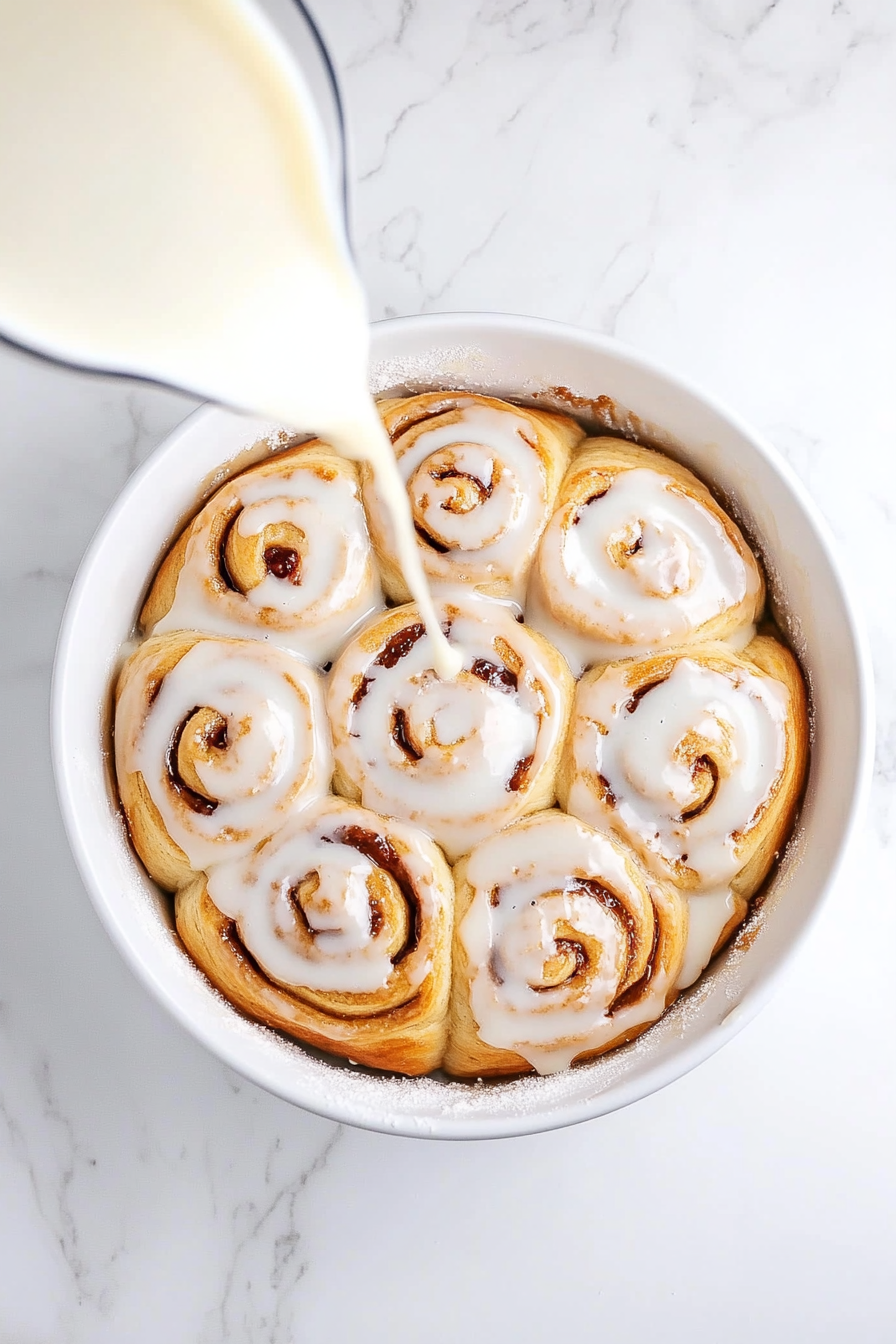 Top-down view of peanut butter and jelly rolls in a pan, with a vanilla glaze being drizzled over the slightly cooled rolls, creating a glossy finish.