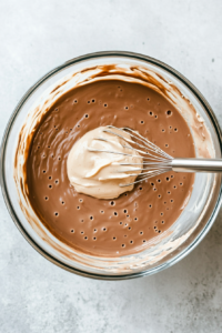 Top-down view of a mixing bowl on a white marble cooktop. Two boxes of chocolate pudding mix and milk are being whisked together, creating a smooth, velvety chocolate pudding mixture.