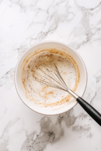 Top-down view of a medium mixing bowl on a white marble countertop. Milk, sugar, beaten eggs, cinnamon, and vanilla are being whisked together into a smooth custard mixture.