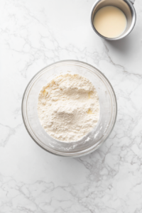 Top-down view of flour, sugar, and salt being mixed in a large bowl with a saucepan of milk and butter cooling beside it, ready for yeast to be added.