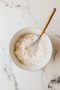 Top-down view of flour, baking powder, and salt being whisked together in a mixing bowl on a clean kitchen countertop, ready to be added to the batter.