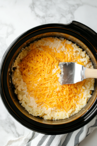 A top-down view of a slow cooker on a clean kitchen countertop with a white marble cooktop background. Inside the slow cooker, a 2-pound package of thawed Southern-style hash brown potatoes, shredded Cheddar cheese, condensed cream of chicken soup, chopped yellow onion, milk, minced garlic, and seasoned salt are being mixed together with a spatula. The scene focuses on the even mixing of the ingredients in the slow cooker.