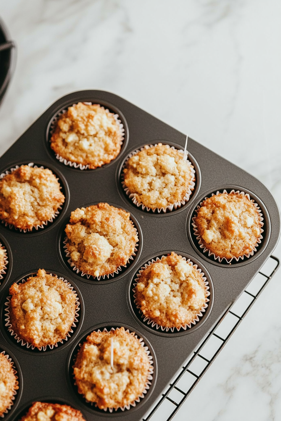 A top-down view of the baked muffins cooling slightly in the muffin tin on a wire rack placed on a clean kitchen countertop with a white marble cooktop background. A toothpick inserted into one muffin comes out clean, and steam is gently rising from the muffins, which are ready to be served.