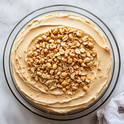 Top-down view of a fully frosted peanut butter cake being garnished with chopped peanuts, adding texture and visual appeal.