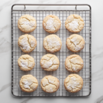 Top-down view of cooled cookies on a wire rack being rolled in confectioners' sugar again for a sweet finish.