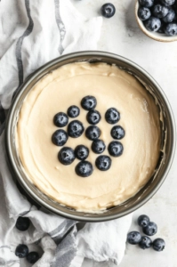 Top-down view of cake batter being poured into a greased and floured 8-inch cake pan, with blueberries visible in the batter, ready to bake.