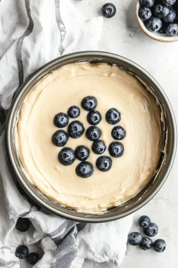 Top-down view of cake batter being poured into a greased and floured 8-inch cake pan, with blueberries visible in the batter, ready to bake.