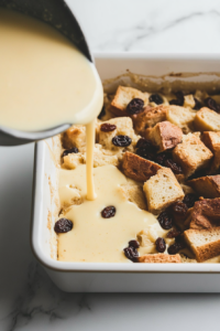 Top-down view of custard being poured over bread pieces and raisins in a baking pan. A fork gently presses down the bread to ensure it absorbs the liquid, on a white marble background.