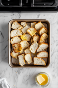 Top-down view of melted butter being drizzled over torn bread pieces in a baking pan, with optional raisins being sprinkled on top. The scene is set on a white marble cooktop background.