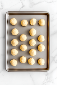 Top-down view of chilled dough being rolled into 1-inch balls and placed on a baking sheet on a white marble cooktop.