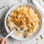 A top-down view of the finished dish being served from the slow cooker onto a plate on a clean kitchen countertop with a white marble cooktop background. The creamy, cheesy hash brown mixture with a crunchy cracker topping is being spooned onto the plate, ready to be enjoyed. The scene focuses on the rich texture and golden topping of the dish, with the plate and serving spoon clearly visible.