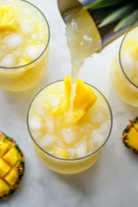 Top-down view of chilled tropical lemonade being poured into glasses filled with ice, garnished with a slice of pineapple or mango, ready to serve on a white marble surface.