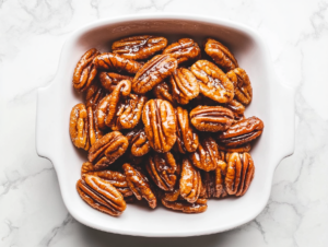 Top-down view of the cooled and hardened candied pecans arranged on a clean white serving dish on the white marble cooktop. The pecans are glossy, sugary, and crunchy, ready to be served as a snack or dessert topping. The scene highlights the shiny texture and golden-brown color of the candied pecans.