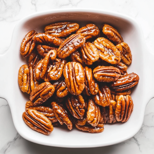 Top-down view of the cooled and hardened candied pecans arranged on a clean white serving dish on the white marble cooktop. The pecans are glossy, sugary, and crunchy, ready to be served as a snack or dessert topping. The scene highlights the shiny texture and golden-brown color of the candied pecans.