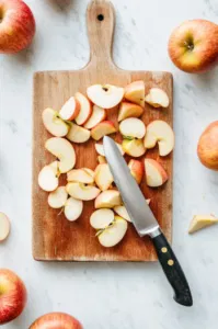 Top-down view of a cutting board with peeled apples being sliced into 1/8-inch thick pieces using a sharp knife, surrounded by apple slices.