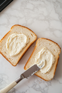 Top-down view of two slices of crusty white bread on a marble countertop. One side of each slice is being spread with mayonnaise using a butter knife.