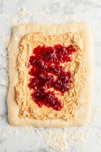 Top-down view of peanut butter and jelly being spread evenly over a rolled-out dough rectangle on a floured surface.