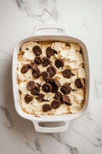 Top-down view of a baking dish with cookie dough pieces on top. Crushed Oreos are being sprinkled evenly over the surface, creating a crunchy topping.