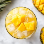 Top-down view of chilled tropical lemonade being poured into glasses filled with ice, garnished with a slice of pineapple or mango, ready to serve on a white marble surface.