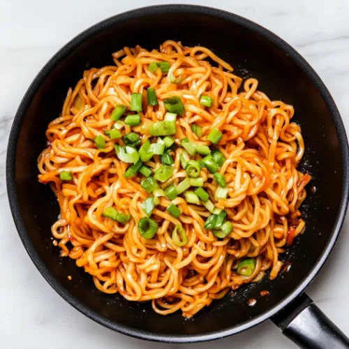 Top-down view of the completed noodle stir-fry in the skillet, garnished with thinly sliced green onions, sitting on a white marble countertop, ready to be served.