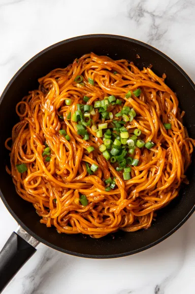 Top-down view of the completed noodle stir-fry in the skillet, garnished with thinly sliced green onions, sitting on a white marble countertop, ready to be served.