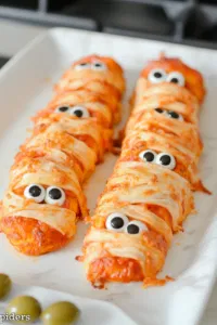 Golden and crispy baked pizza mummies on the white marble cooktop. Each mummy is topped with two edible spooky eyes, and olive 'spiders' are arranged around the plate for a spooky effect. Optionally dusted with flour for a more mummy-like look.