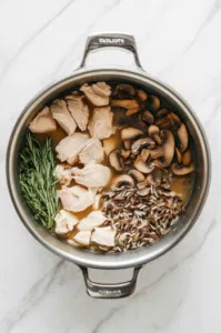 [This image shows the stockpot with a mixture of chicken stock being poured in, alongside chicken breasts, diced mushrooms, wild rice, rosemary, and bay leaves.]