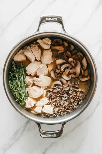 [This image shows the stockpot with a mixture of chicken stock being poured in, alongside chicken breasts, diced mushrooms, wild rice, rosemary, and bay leaves.]