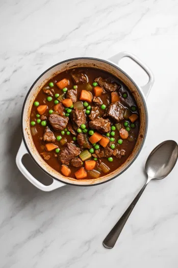 This image shows a bowl of hearty beef stew, filled with tender beef chunks, carrots, potatoes, celery, and peas, served warm with fresh herbs as garnish