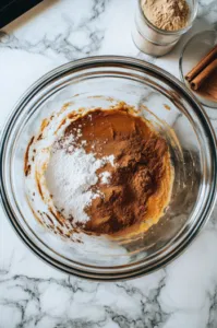 Vanilla extract, sour cream, and a pinch of salt are added to the cream cheese mixture in the glass bowl on the white marble cooktop. A spatula is nearby for scraping the sides of the bowl as the ingredients combine.