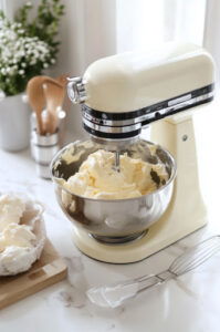 This image shows softened cream cheese and packed brown sugar being beaten on medium speed in a stand mixer until light and fluffy, with the bowl being scraped as needed.