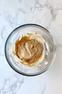 A glass mixing bowl on the white marble cooktop contains shortening, peanut butter, brown sugar, and white sugar being beaten together until smooth. The creamy mixture is ready for the next ingredients