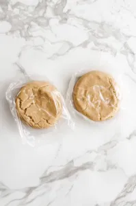 The cookie dough divided into two portions, shaped into disks, and wrapped in plastic wrap on the white marble cooktop. The disks are ready to be refrigerated for 1 hour to firm up.