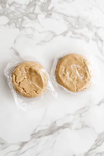 The cookie dough divided into two portions, shaped into disks, and wrapped in plastic wrap on the white marble cooktop. The disks are ready to be refrigerated for 1 hour to firm up.