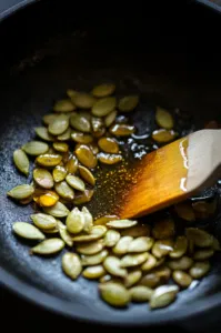 This image shows a spatula stirring honey into sautéed pumpkin seeds in a skillet, ensuring each seed is well coated.