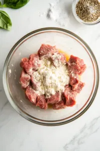 This image shows cubed beef being tossed in a bowl with a mixture of flour, garlic powder, salt, and pepper to ensure an even coating.