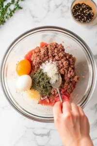 A glass bowl on the white marble cooktop filled with ground beef, salt, black pepper, thyme, egg, and breadcrumbs. Hands are mixing the ingredients thoroughly to form the burger patty mixture.