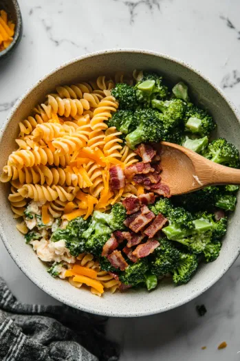 This image shows cooked pasta, steamed baby broccoli florets, diced bacon, and cheddar cheese being added to the bowl with the dressing, ready to be mixed.