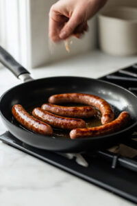 This image shows bratwurst being added to a hot pan with oil, cooking until browned on all sides.