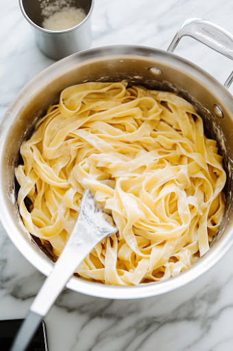 This image shows fettuccine being boiled in a pot of water, with steam rising. A cup of the cooking water is being reserved for later use.