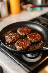 A black skillet on the white marble cooktop with burger patties sizzling in cooking oil. The patties are developing a golden-brown crust as they cook for 3-4 minutes on each side.