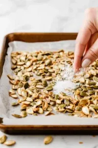 This image shows roasted pumpkin seeds on a parchment-lined baking sheet, being sprinkled with flaky sea salt for flavor.]