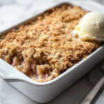 This image shows a warm serving of fruit crumble in a bowl, topped with a scoop of vanilla ice cream. The ice cream is slightly melting over the golden, crispy topping