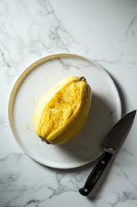 This image shows a cooled spaghetti squash being cut in half lengthwise, revealing its soft, stringy flesh inside.]