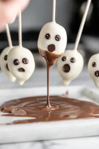 Ghost-shaped cake pops are being removed from their molds on the white marble cooktop. Each pop is being dipped into the smooth white candy melt, with the excess dripping off before being placed on parchment paper.