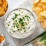 This image shows a white bowl filled with spicy cottage cheese dip, drizzled with chili oil and garnished with fresh scallions and garlic chives, ready to serve.