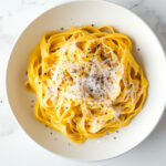 This image shows the finished Pumpkin Alfredo Pasta in a white bowl, topped with extra Parmesan cheese and seasoned to taste, ready to be served.