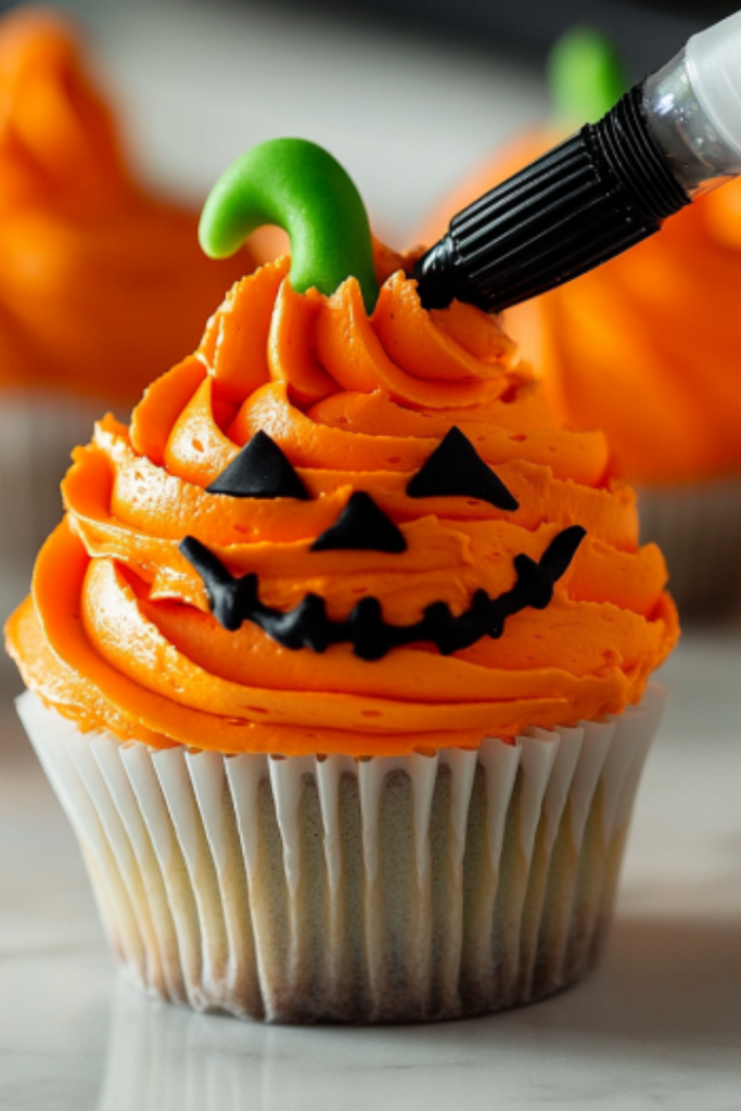 Orange buttercream is piped in bands from the bottom to the top of a cupcake on the white marble cooktop. A green stem and black face are added for a pumpkin design.