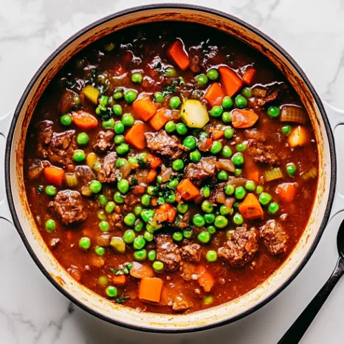 This image shows a bowl of hearty beef stew, filled with tender beef chunks, carrots, potatoes, celery, and peas, served warm with fresh herbs as garnish