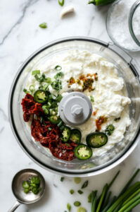 This image shows a food processor filled with cottage cheese, pickled jalapenos, sun-dried tomatoes, salt, scallions, and garlic chives, about to be blended into a smooth dip.